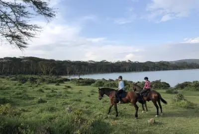 Two riders on horseback crossing acacia grassland beside Lake Naivasha
