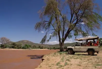 Safari vehicle parked under an acacia beside a red Samburu riverbed
